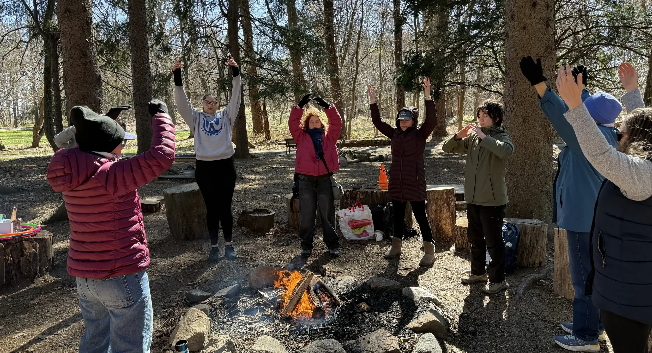 Women gathered around a fire with their arms raised in a wooded clearing